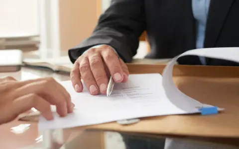 Dwie osoby przy biurku omawiają dokumenty. Jedna z nich, w garniturze, wskazuje długopisem na tekst. To może być poświadczenie notarialne przy wynajmie mieszkania.