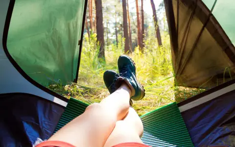 Nogi w butach trekkingowych odpoczywają w namiocie. Słońce przebija się przez drzewa. Idealne miejsce, by zanocuj w lesie.