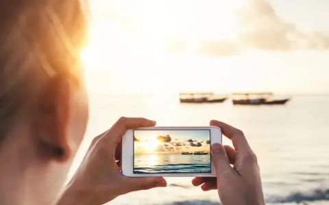 Une personne prend une photo d'un coucher de soleil sur la mer avec des bateaux au loin. Une belle photo à faire à la plage.