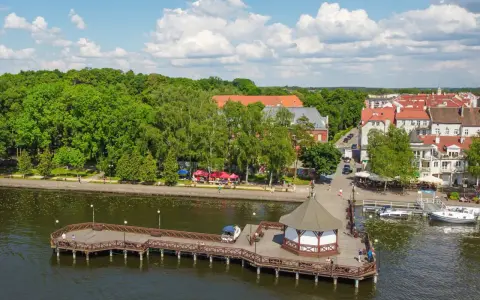 Malowniczy widok na mazury: drewniane molo z altaną, jezioro, zielone drzewa i urokliwe miasteczko z czerwonymi dachami.