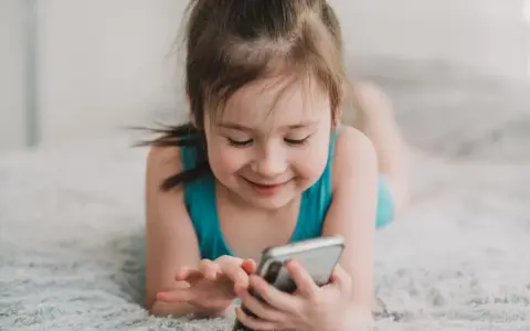 Uśmiechnięta dziewczynka leży na dywanie, bawiąc się telefonem. Wpływ telefonu komórkowego na mózg dziecka jest tematem dyskusji.