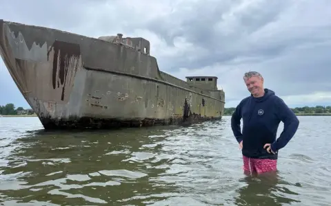 Mann steht im Wasser neben einem verwitterten Betonschiff in Wismar.