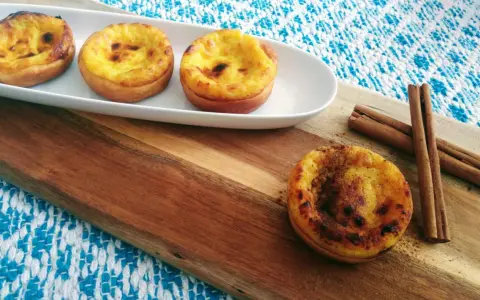 Vier goldbraune, karamellisierte Pastéis de Nata auf einem weißen Teller, daneben Zimtstangen auf einem Holzbrett.