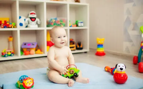 Uśmiechnięte, 8-miesięczne dziecko nie siedzi, bawi się zielonym autkiem na niebieskim kocyku. W tle regał z zabawkami.