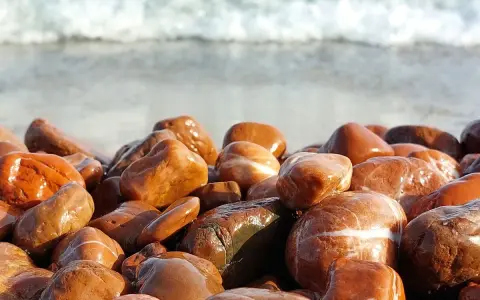 Gładkie, mokre kamienie na plaży, przypominające jaspis czerwony właściwości. W tle widać falę.