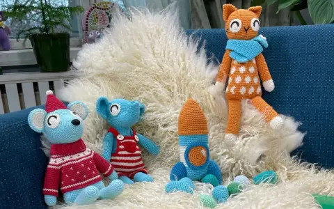 En virkad amigurumi katt med orange och vita prickar sitter på en lurvig matta bredvid en blå mus med tomteluva och en blå elefant i hängselbyxor.