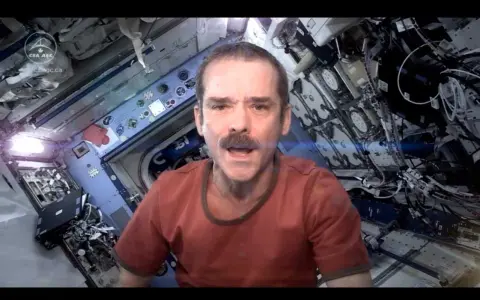 Chris Hadfield singt "Space Oddity" in der ISS. Sein Gesicht mit Schnurrbart ist im Vordergrund, der Raumschiff-Innenraum im Hintergrund.