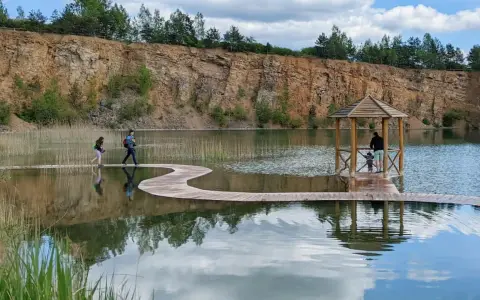 Malownicza trasa Gródka Jaworzno z drewnianym molo i altanką. Dwie osoby spacerują po falistej kładce nad wodą.