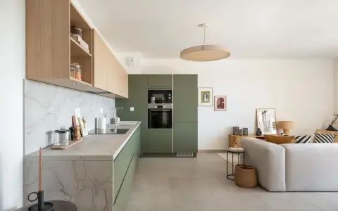 Cuisine moderne avec des armoires vert olive, un plan de travail en marbre et des étagères en bois clair. Un salon adjacent est visible.