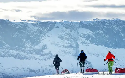 Tre skidåkare med slädar drar sig fram över snöfältet mot ett snötäckt berg. En fantastisk upplevelse att besöka Jokkmokk.