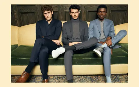 Three men in suits sit on a couch. The man on the left wears dark dress pants, while the other two wear suit pants.