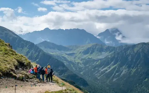 Grupa turystów odpoczywa na szlaku z widokiem na majestatyczne Tatry. W oddali majaczy Kasprowy Wierch, otoczony chmurami.