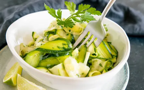 Frischer Zucchini-Salat mit Petersilie und Limettenspalten, serviert in einer weißen Schüssel. Eine Gabel steckt im Salat.