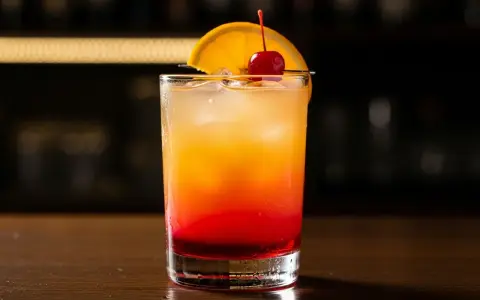 En klassisk tequila sunrise med lager av rött och orange, garnerad med apelsinskiva och körsbär.