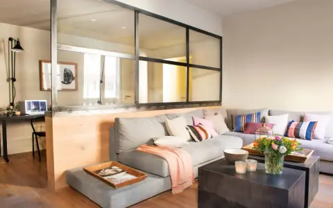 Un salón moderno con una pared de cristal que separa un área de trabajo. Un sofá gris con cojines coloridos y una manta rosa.