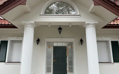 Elegancki pilaster zdobi fasadę domu, wspierając dach nad wejściem. Obok okna i drzwi, tworzy klasyczną kompozycję.