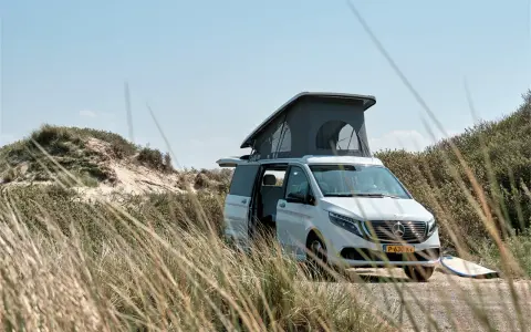Vit el campervan med uppfällt tak står parkerad vid en sanddyn. En surfbräda ligger bredvid.