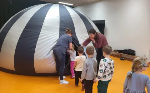 Dzieci i opiekunki wchodzą do dmuchanej kopuły, która przypomina planetarium. To miejsce, gdzie można oglądać filmy z przesłaniem, odkrywając tajemnice wszechświata.