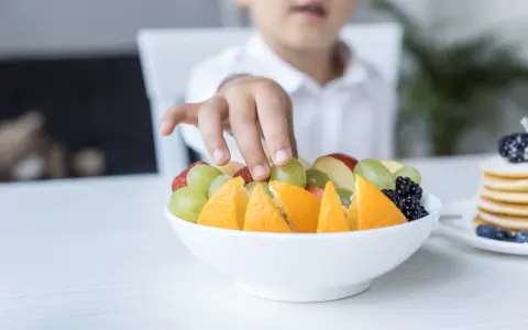 Małe rączki sięgają po miskę pełną owoców: pomarańczy, truskawek, jabłek i winogron dla niemowlaka.