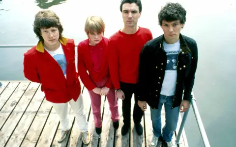Die Band Talking Heads steht auf einem Holzsteg. David Byrne trägt ein rotes Hemd, Tina Weymouth ein pinkes Outfit.