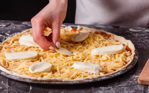 Dłoń układa kawałki sera na pizzy, która jest już posypana startym żółtym serem.