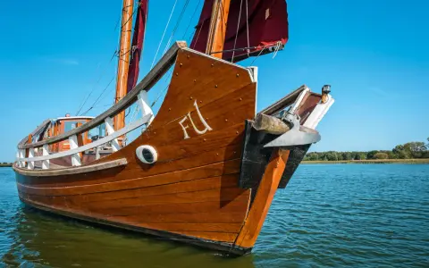 Ein hölzernes Dschunke Schiff gleitet auf ruhigem Wasser unter blauem Himmel. Die Segel sind teilweise gesetzt.