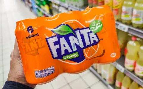 Opakowanie 6 puszek Fanty Orange w ręce, na tle sklepowych półek z napojami.