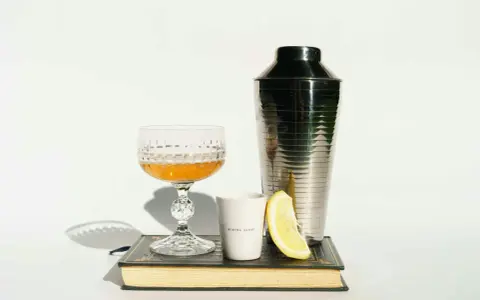Szklanka z drinkiem, shaker, cytryna i książka. Idealne akcesoria, by dowiedzieć się, z czym pić amarula.
