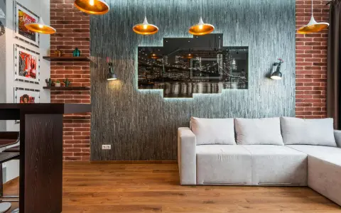 Nowoczesny salon w stylu loft: szara kanapa, ceglana ściana, panel z nocnym widokiem miasta i złote lampy.