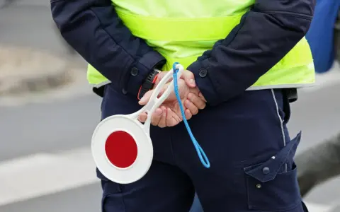Policjant w kamizelce odblaskowej trzyma za plecami lizak do zatrzymywania pojazdów.