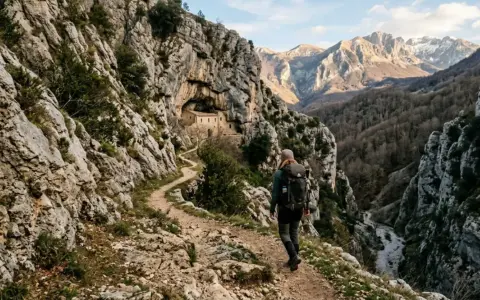 Un escursionista percorre un sentiero roccioso verso un eremo incastonato nella roccia, un'avventura nei cammini in Abruzzo.