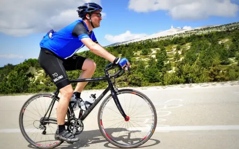 Ciclista in maglia blu affronta la **salita** del **Mont Ventoux**, con la cima innevata in lontananza.