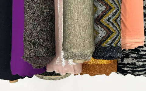 Rouleaux de tissus aux textures variées : noir uni, violet, motifs complexes, rose pâle, beige chiné, zigzags multicolores, pêche, et noir et blanc. Une matière mesh se distingue par sa finesse.