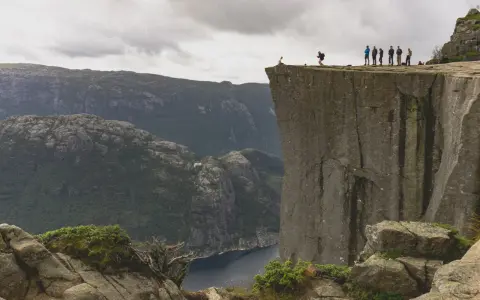 Grupa ludzi na krawędzi klifu w **Stavanger, Norwegia**, z widokiem na fiord i góry.