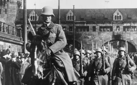 Soldaten marschieren in Formation, ein Reiter führt sie an. Ein historisches Bild der Rheinlandbesetzung 1936.