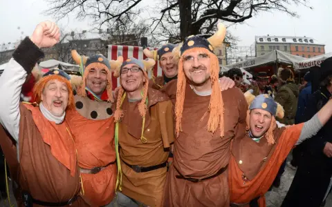 Fröhliche Männer in historischer Wikinger-Kleidung mit Hörnerhelmen und orangefarbenen Perücken feiern ausgelassen.