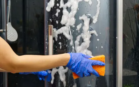 Main bleue, éponge orange pour nettoyer paroi douche. Mousse et bulles sur la paroi vitrée.