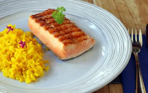 Un delicioso salmón a la plancha con marcas de parrilla, servido con arroz amarillo y decorado con perejil y flores moradas.