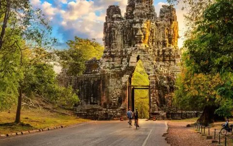 Two cyclists approach a massive stone gateway, one of the ancient Cambodia temples, bathed in golden sunlight.