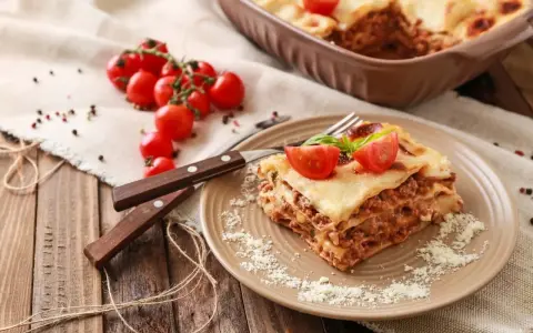 Pyszna, wielowarstwowa lasagne z mięsem mielonym, polana sosem beszamelowym i posypana parmezanem. Na talerzu ozdobiona pomidorkami koktajlowymi i bazylią.