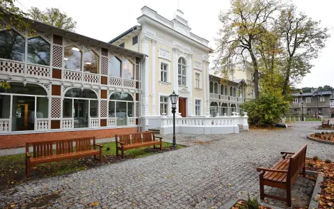 Elegancki budynek z ażurowymi werandami, przypominający dawny **Hevelianum dom zdrojowy**. Brukowany dziedziniec z ławkami i jesiennymi liśćmi.
