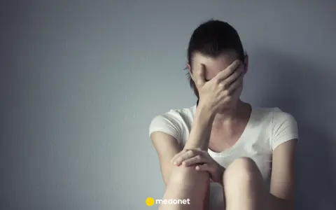 Kobieta w białej koszulce zakrywa twarz dłońmi, siedząc przy szarej ścianie. Może to być symbol smutku lub problemów, z którymi zmaga się, jak np. depresja, którą czasem leczy się lekiem lerivon.