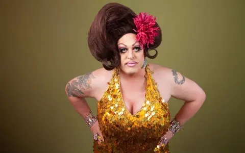Osoba w złotej, cekinowej sukience i peruce, z kwiatem we włosach. To przykład tego, co to jest drag queen – artystyczna forma ekspresji.
