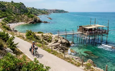 Ciclista lungo la via dei trabocchi, con un trabocco in legno sull'acqua cristallina e una spiaggia rocciosa.