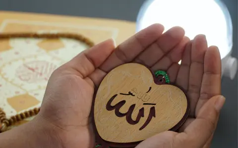 Hände halten ein Herz mit dem Wort "Allah" in arabischer Schrift, ein Symbol für ikhlas sure. Gebetskette und ein Buch liegen im Hintergrund.