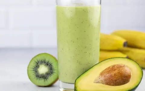 Zielony koktajl z awokado, kiwi i bananów w szklance, obok połówka awokado i kiwi.