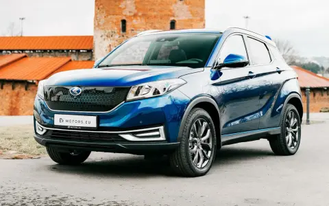Błękitny SUV EV Motors.eu z charakterystycznym grillem, zaparkowany przed zabytkowym budynkiem z cegły.
