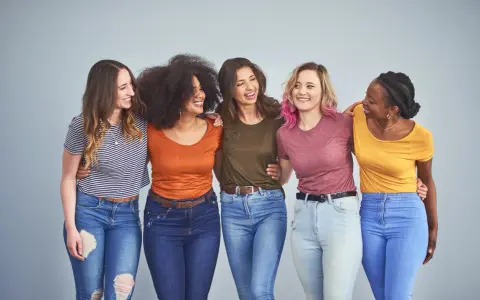 Cinque amiche sorridono, vestite con jeans e magliette colorate, tra cui spiccano tonalità marrone e bianco.