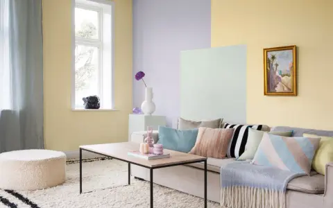 Ett vardagsrum med pastell inredning. Ljusgula, ljuslila och ljusgröna väggar skapar en mjuk atmosfär. En beigefärgad soffa är fylld med kuddar i olika pastellfärger och mönster.