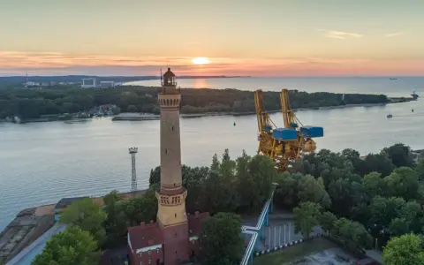 Zachód słońca nad morzem, obok najwyższej latarni morskiej w Polsce, widać też portowe dźwigi.